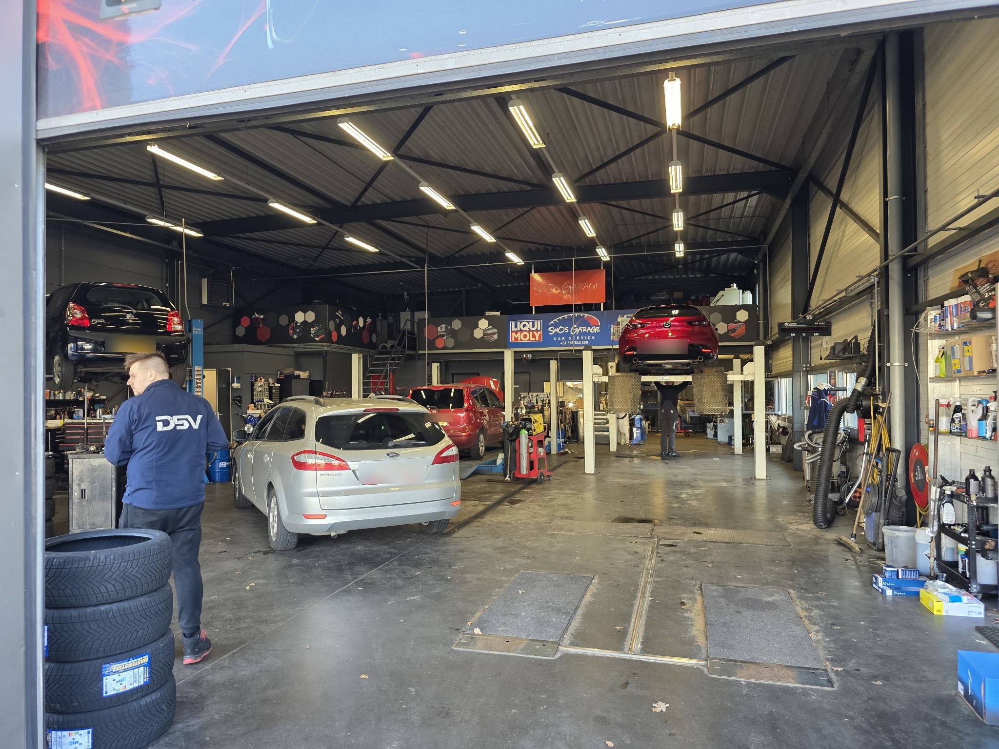 Binnen in de werkplaats van SwOs Garage, met auto's op hefbruggen en een monteur aan het werk